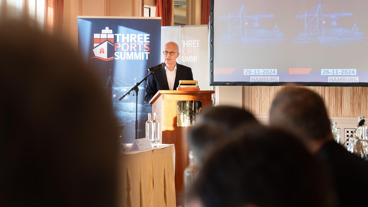 man die toespraak geeft achter een desk voor een zaal met toehoorders, achter hem staan roll-up banners van three ports summit